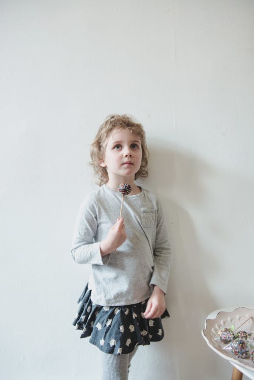 Free Content kid in skirt with rabbits standing near dish with desserts against white wall with delicious cake pop in hand and looking away dreamily Stock Photo