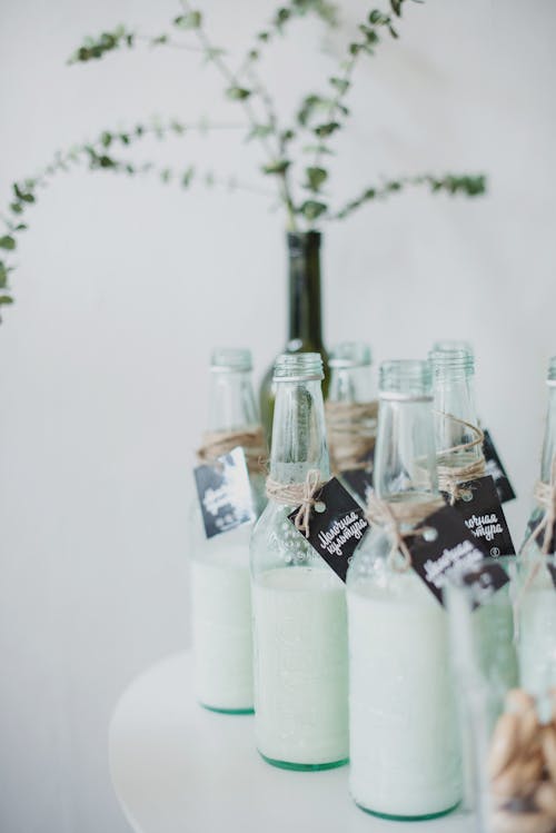 Free Glass bottle of milk with tied labels placed on white round table near vine bottle with green tree branch illustrating spring and Easter concept Stock Photo