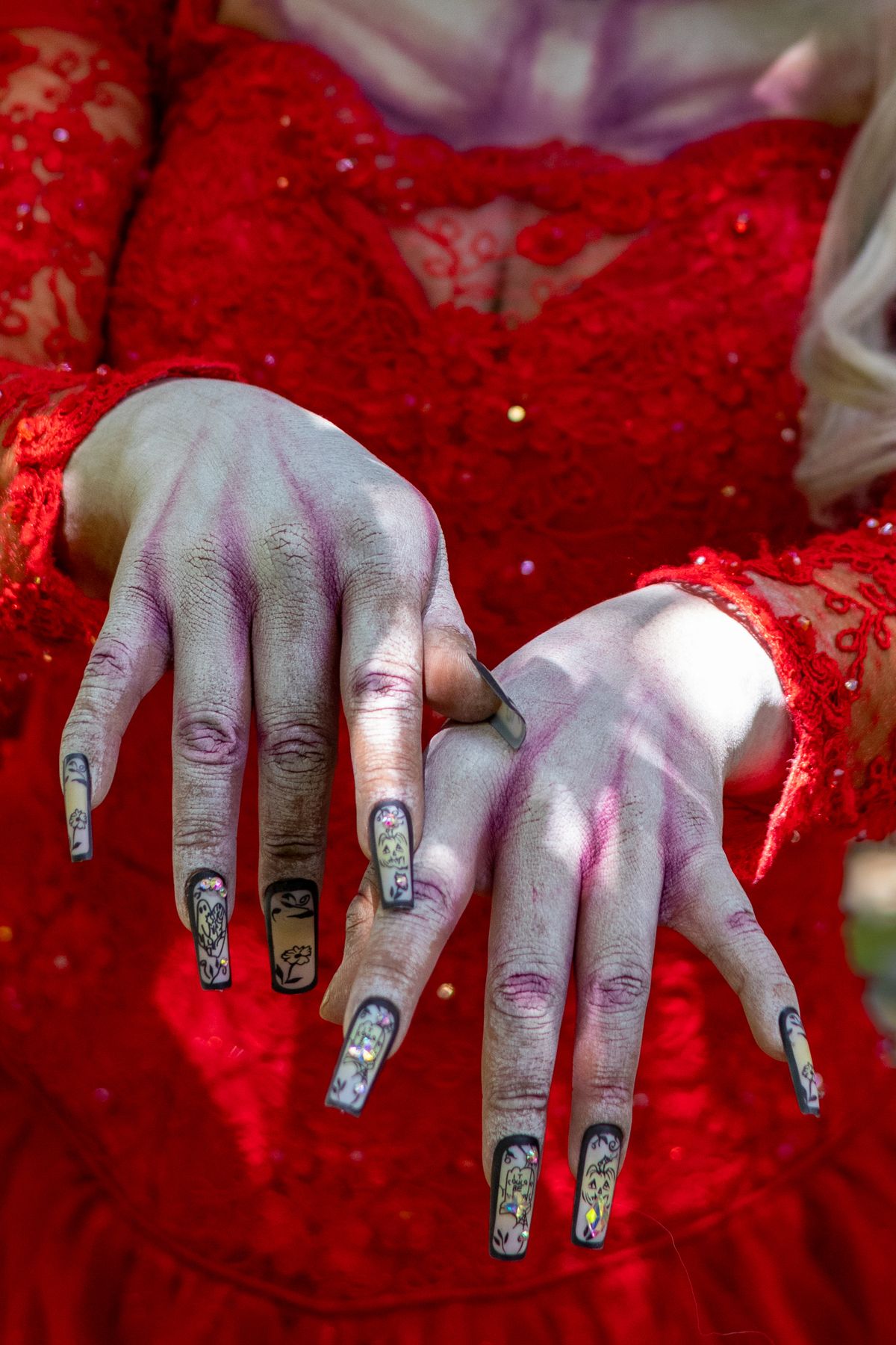 Close-up of zombie-themed costume with gothic nail art and red lace dress, shot outdoors in daylight.