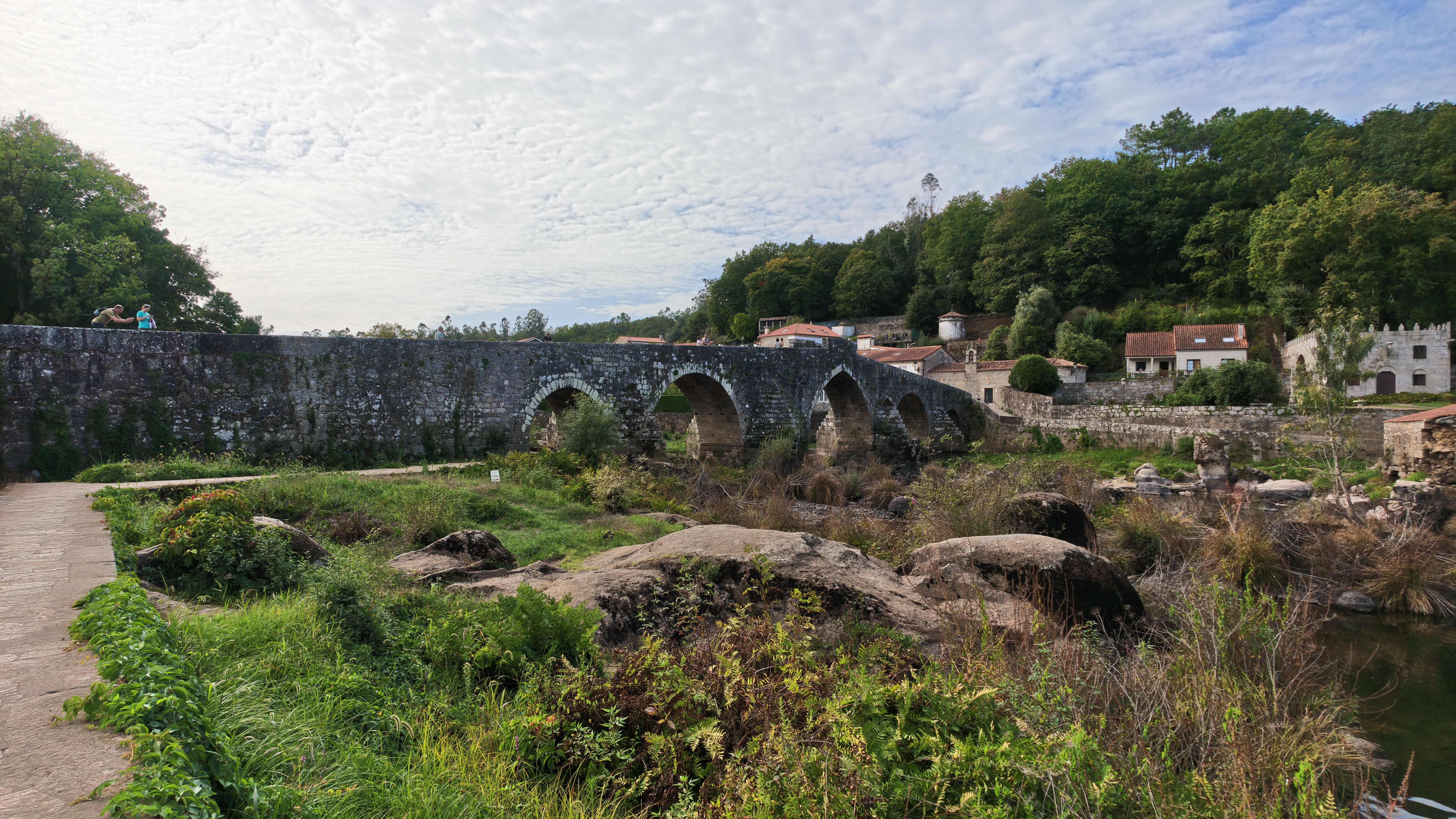 Ancient stone arch bridge with lush greenery in a rural village setting on a bright day.