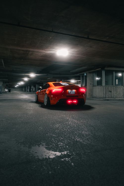 Free A sleek orange sports car parked in a deserted underground garage, illuminated by dim overhead lighting. Stock Photo