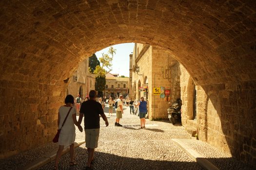 A sunny day scene with tourists walking under a stone archway in a historic town.