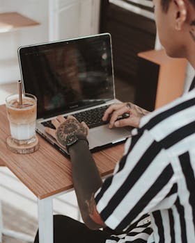A tattooed individual working on a laptop in a café setting, enjoying a coffee drink.