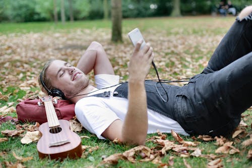 Man in White Shirt Lying on Green Grass Field Holding White Smartphone