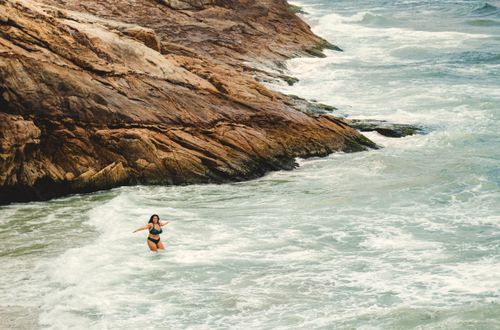 Free A woman in a bikini wading through ocean waves near a rocky shoreline, enjoying the natural scenery. Stock Photo