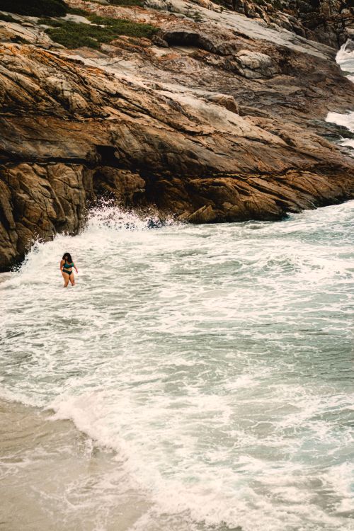 Free A woman stands near rocky cliffs by the sea, embraced by ocean waves, showcasing adventure and natural beauty. Stock Photo