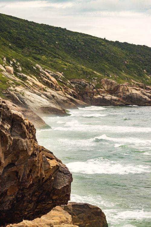Free Dramatic rocks and lush green hills meet the ocean on a cloudy day. Stock Photo