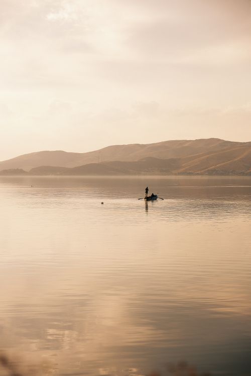 Free Peaceful scene of a lone fisherman on a calm lake at sunrise, surrounded by soft mountains. Stock Photo