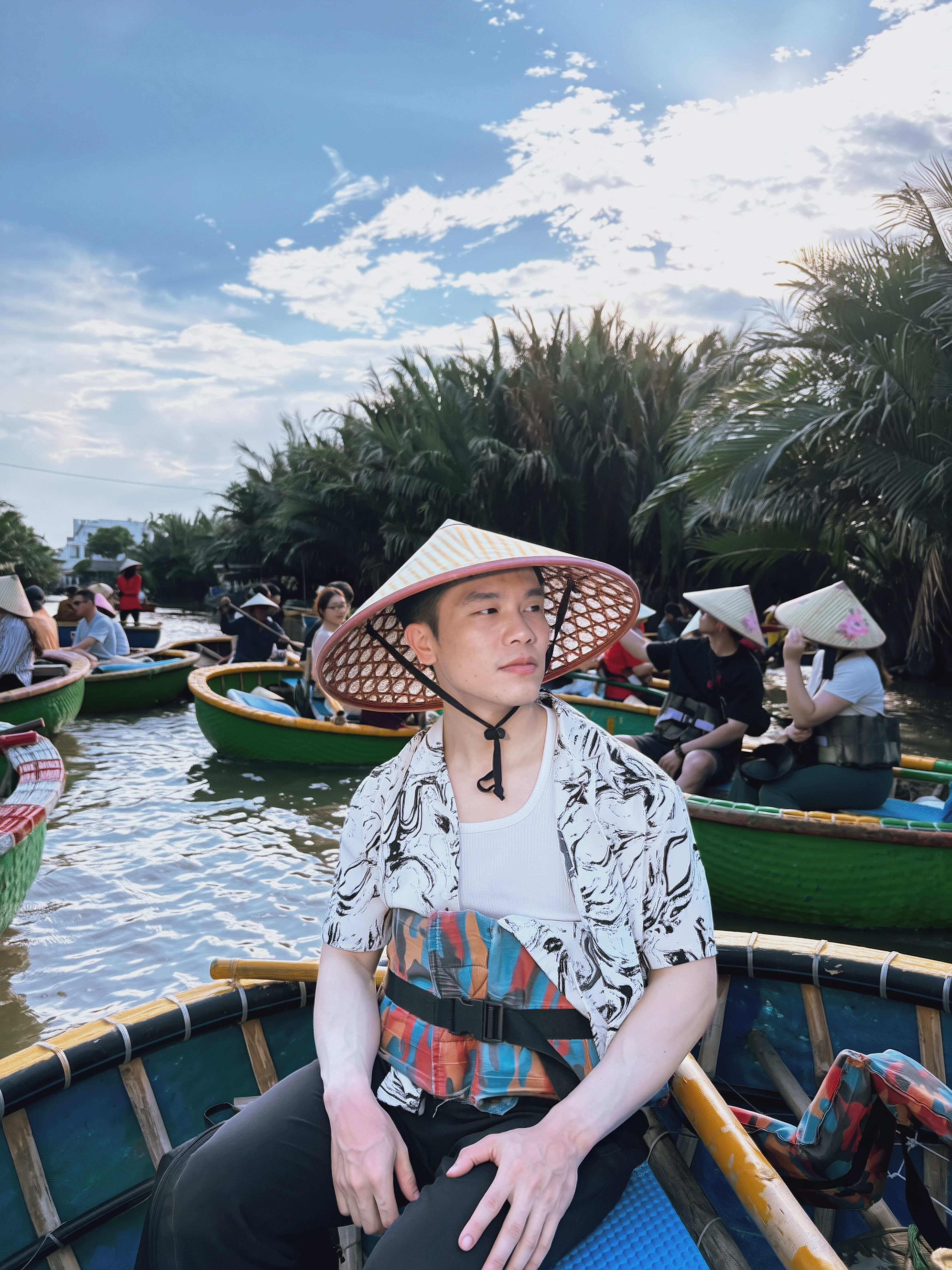 A man in traditional Vietnamese straw hat sits on a bamboo basket boat amidst tropical scenery.