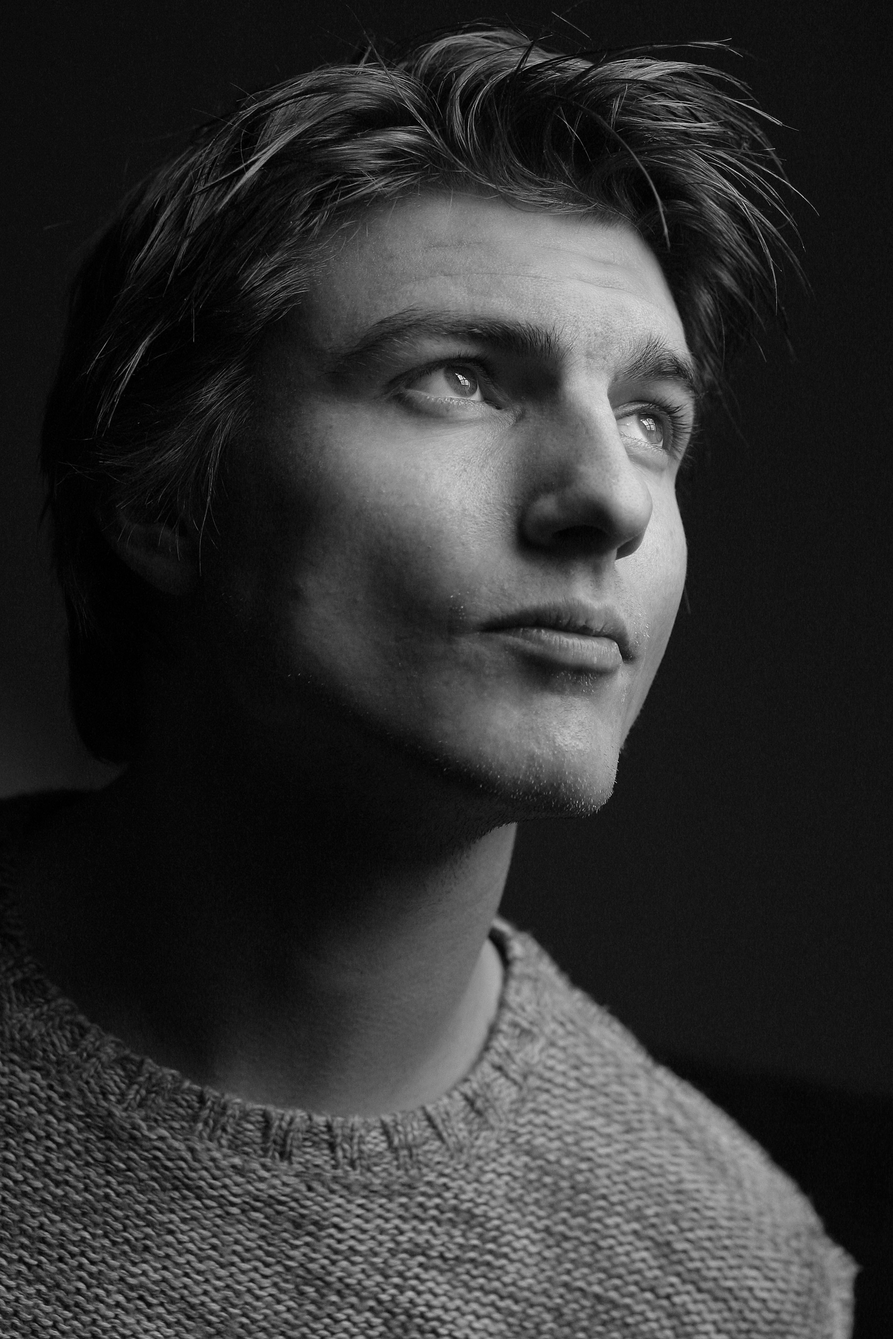 Black and white portrait of a young man in a studio setting, wearing knitwear and looking upwards pensively.