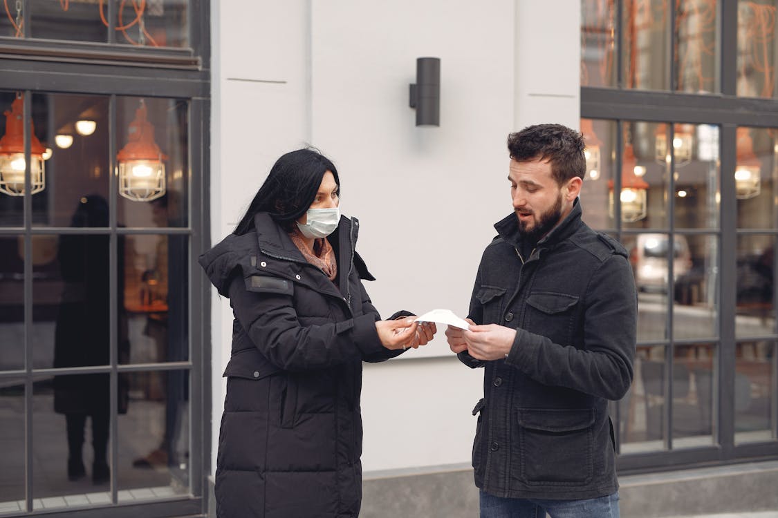 Careful female volunteer in black down jacket suggesting protective facial mask for bearded male pedestrian on city street in cold season during coronavirus pandemic