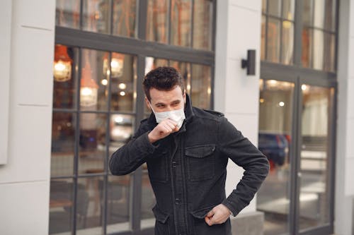 Unhealthy male wearing warm jacket and protective facial mask coughing while standing alone on urban street against city restaurant exterior in cold season