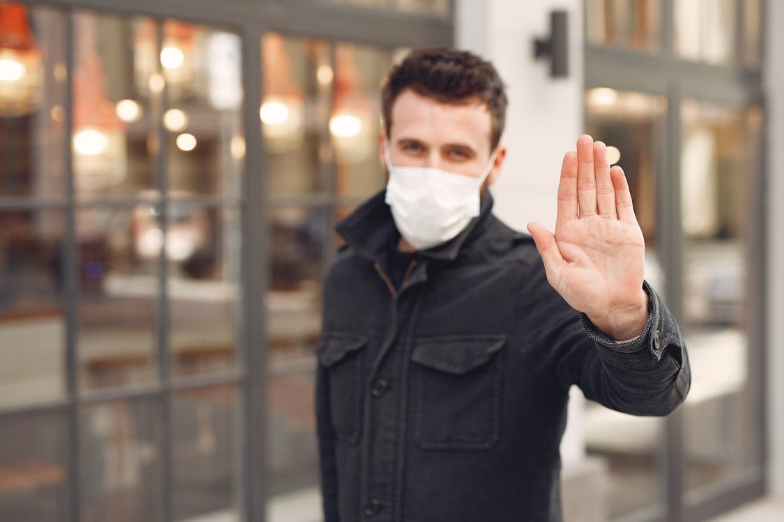 Worried male wearing warm jacket and protective facial mask making prohibitory gesture to stop from social distancing breaking while standing on street against city building exterior in cold season
