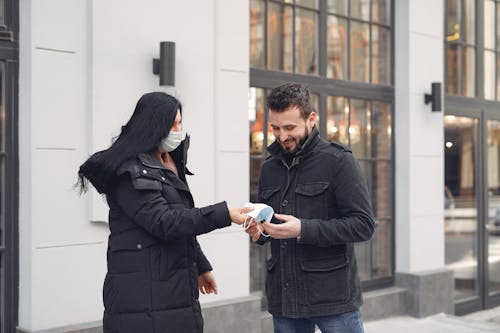 Cheerful bearded man taking protective facial mask from girlfriend while standing together against city building facade during coronavirus pandemic at daytime
