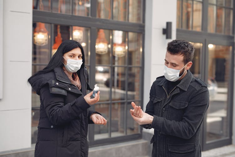 Young Couple In Medical Masks Using Sanitizer Gel Against Urban Building Facade