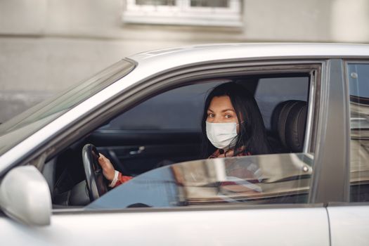 Female driver wearing casual outfit and protective mask on face sitting in automobile with opened window during Coronavirus pandemic near city building and looking at camera