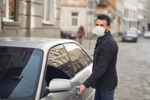 Bearded male driver wearing warm jacket and protective facial mask opening door of auto on blurred city background during Coronavirus pandemic and looking at camera