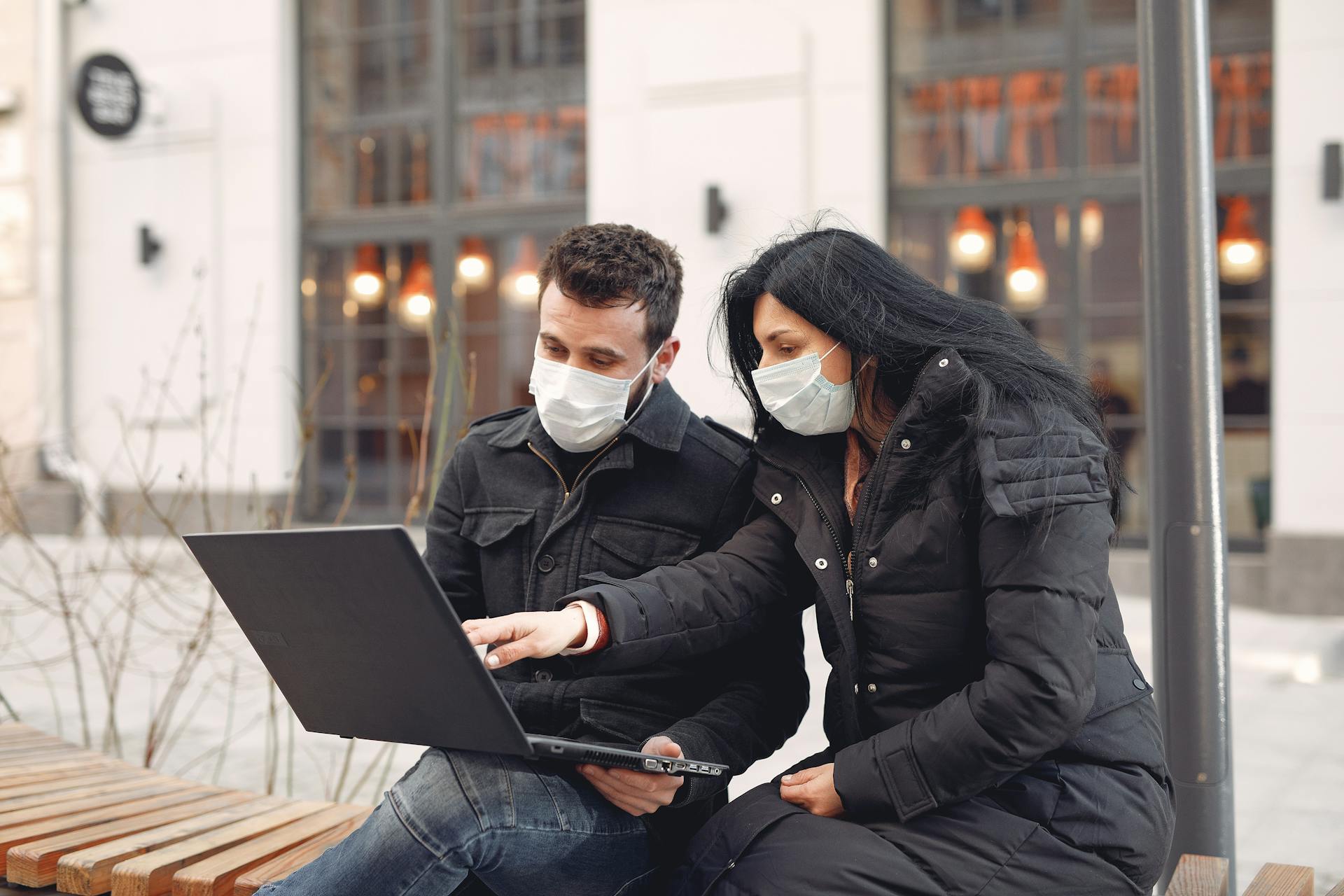 Focused colleagues wearing warm outerwear with protective masks on faces surfing laptop during remote teamwork sitting on wooden bench against urban building