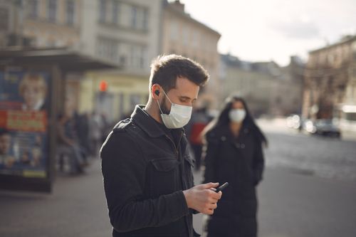 Free Side view of young male wearing warm jacket and protective facial mask surfing mobile phone not far from bus stop on city street during coronavirus pandemic Stock Photo