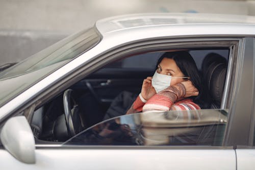 Female driver wearing warm outfit adjusting protective facial mask while sitting in auto with opened window during coronavirus pandemic in city and looking at camera