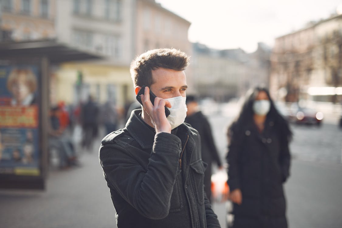 Young man in medical mask speaking on smartphone on urban street during coronavirus pandemic
