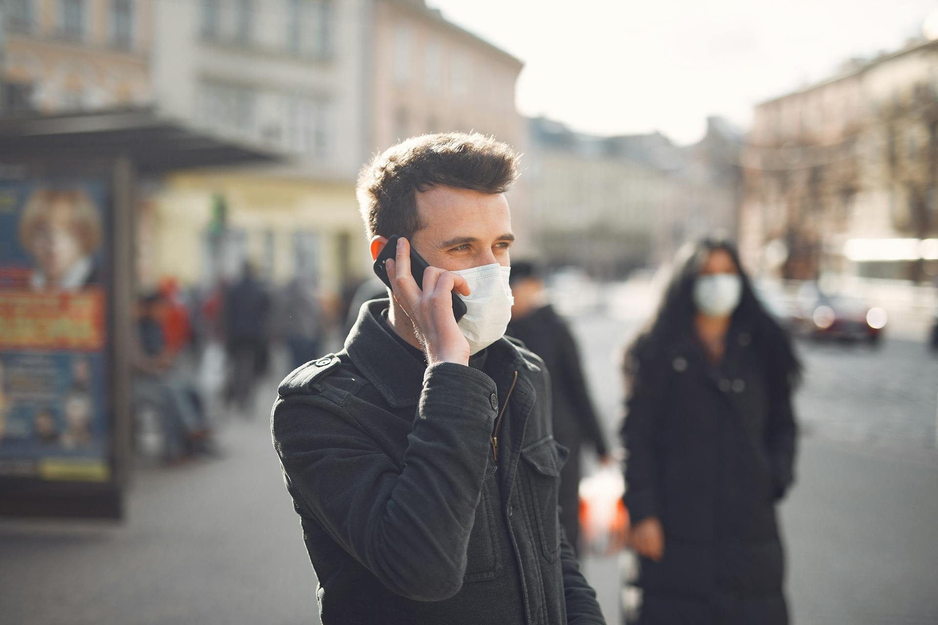 Attentive male wearing warm jacket and protective facial mask standing not far from bus stop during phone call on city street in cold season