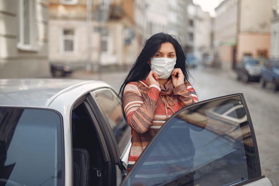 Woman in striped sweater adjusting mask while exiting car on city street during daytime.
