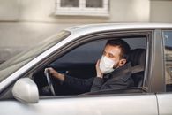 Serious bearded male driver wearing warm jacket and protective facial mask using wireless earphones sitting in auto with opened window during coronavirus pandemic