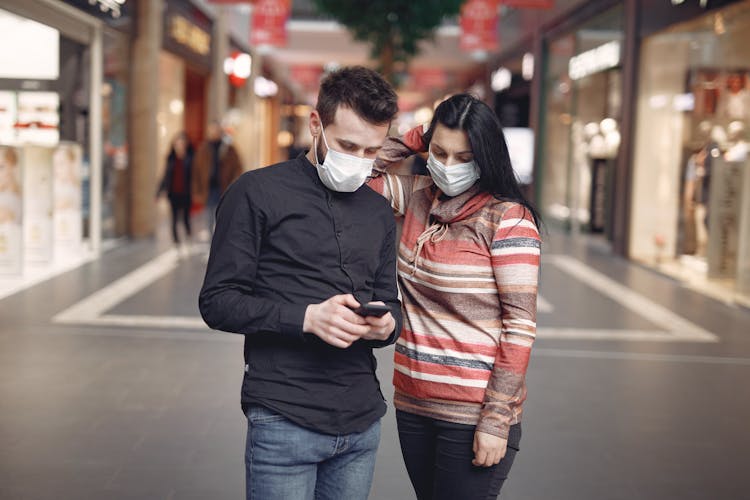 Focused Young Couple Wearing Medical Masks With Smartphone In Shopping Center