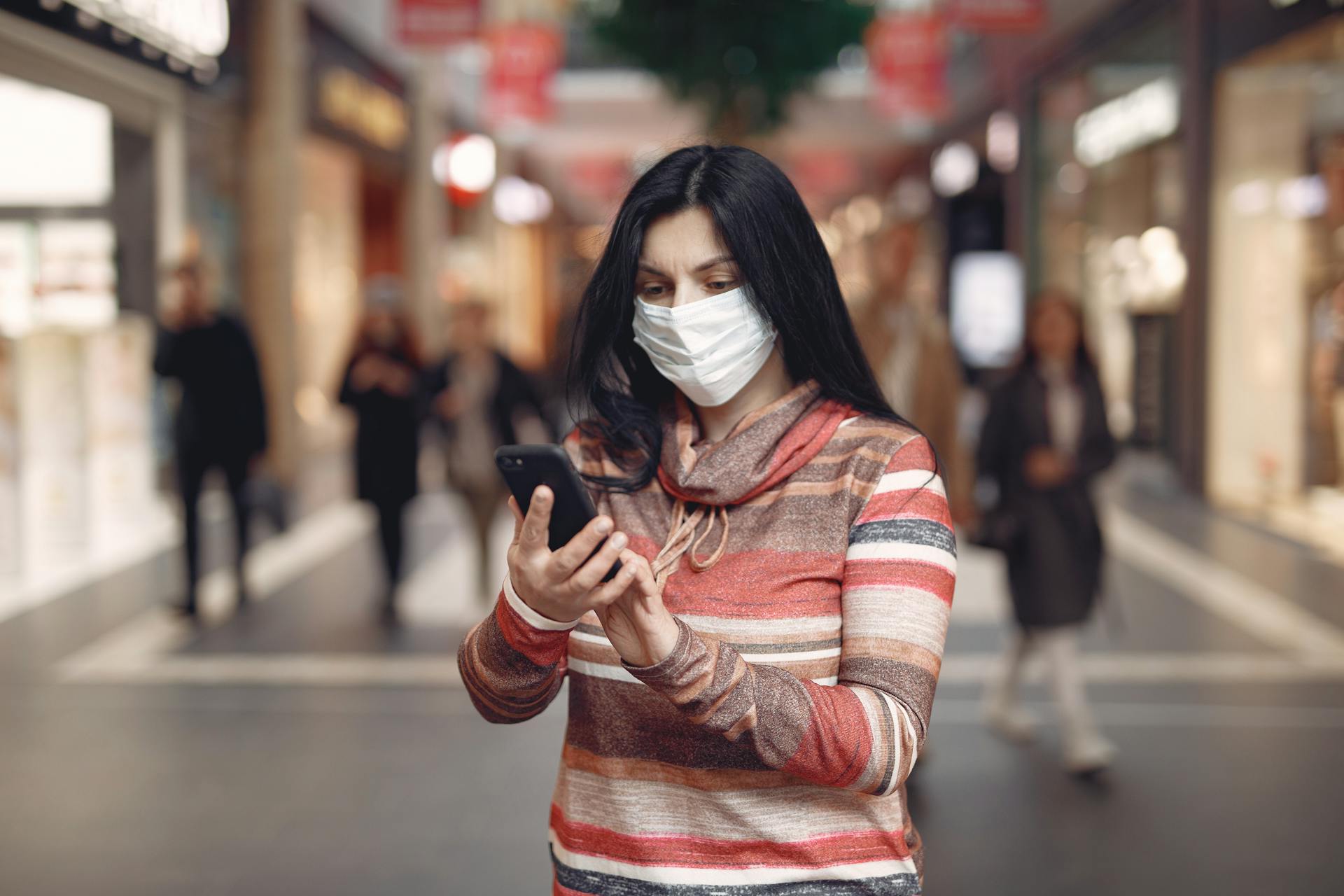 Young female customer in striped wear and protection mask on face using smartphone while standing in shopping mall during coronavirus pandemic