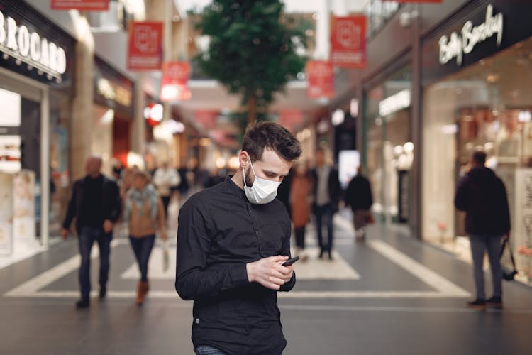 Young Man In Disposable Mask Using Smartphone In Middle Of Shopping Center