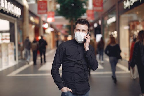 Stylish male customer speaking on smartphone through disposable mask while walking near modern stores