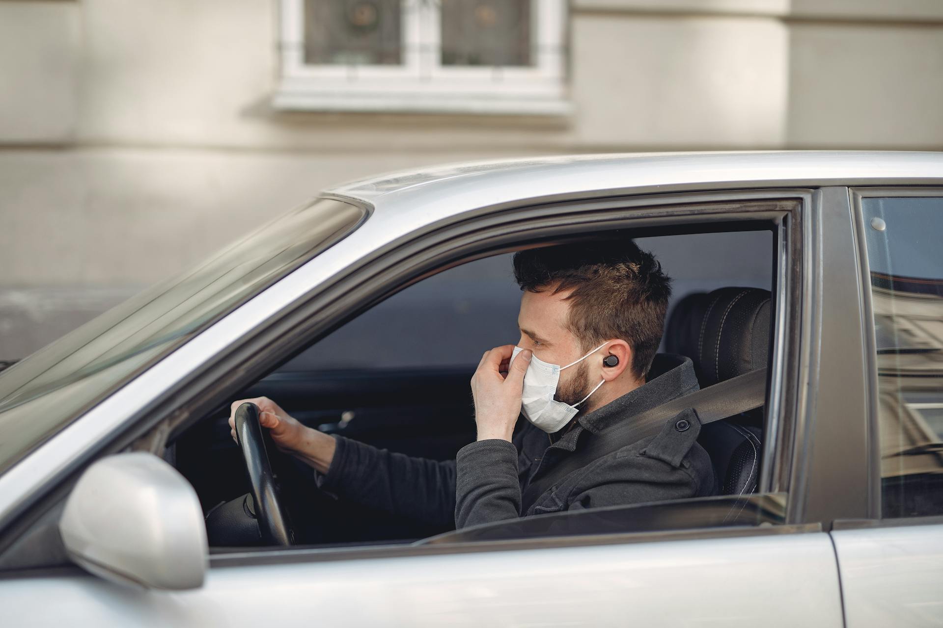 Serious man in disposable mask and earbuds driving car at daytime
