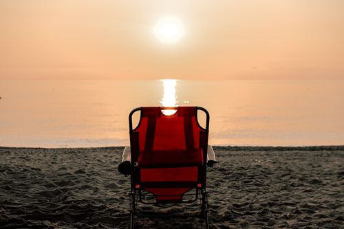 Free A solitary red chair on the beach facing a calm sunset over the water. Stock Photo