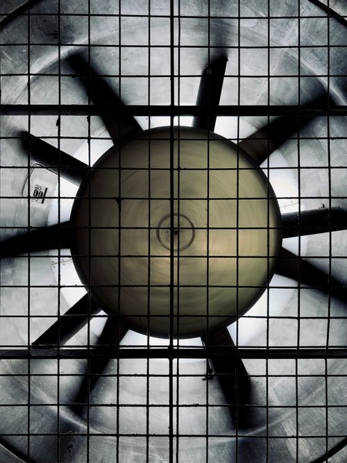 Free Top-down view of a large industrial ceiling fan through a grid, showcasing its metallic blades. Stock Photo