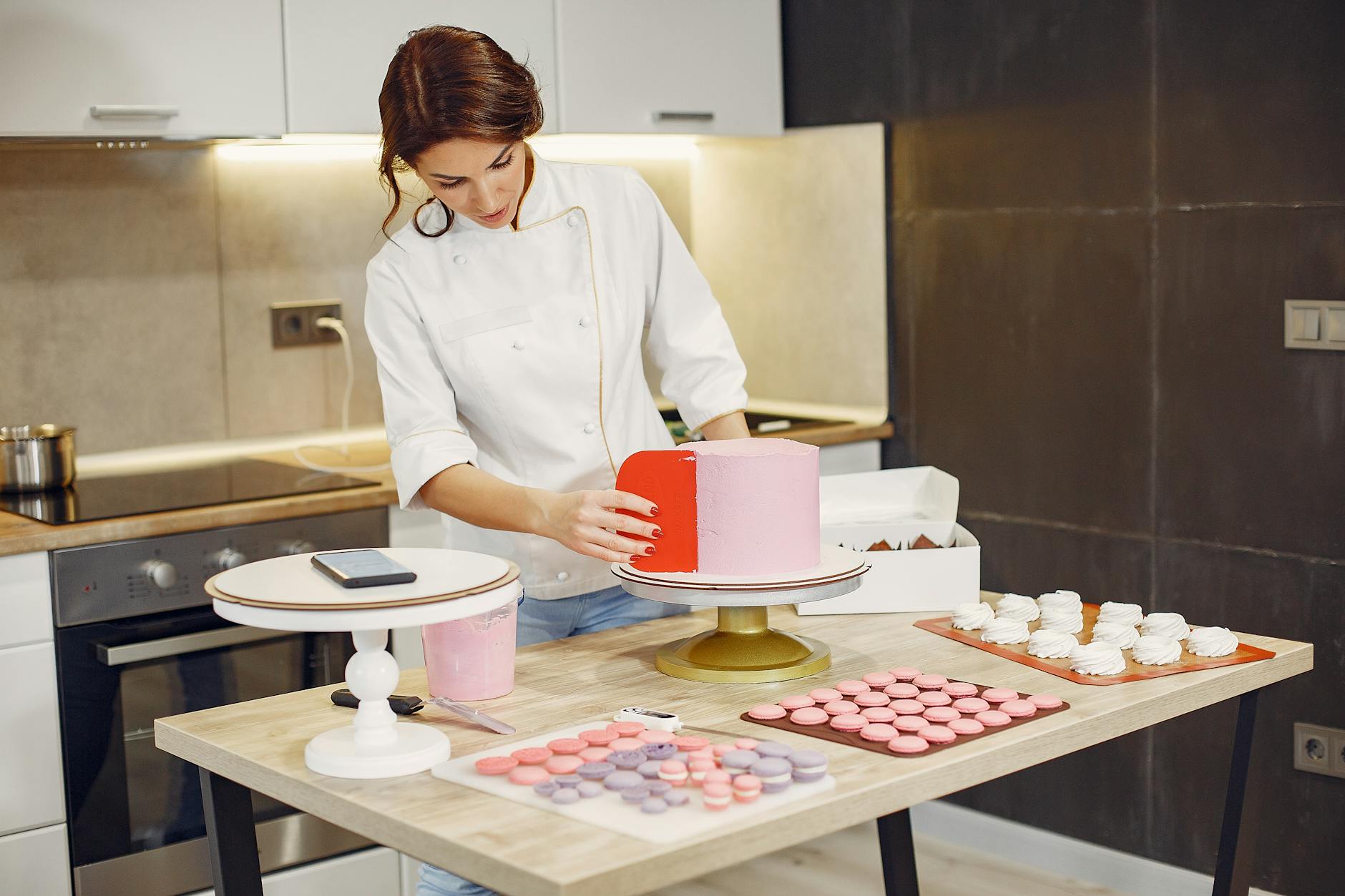 Focused female pastry chef preparing cake and desserts with smartphone aside in modern kitchen