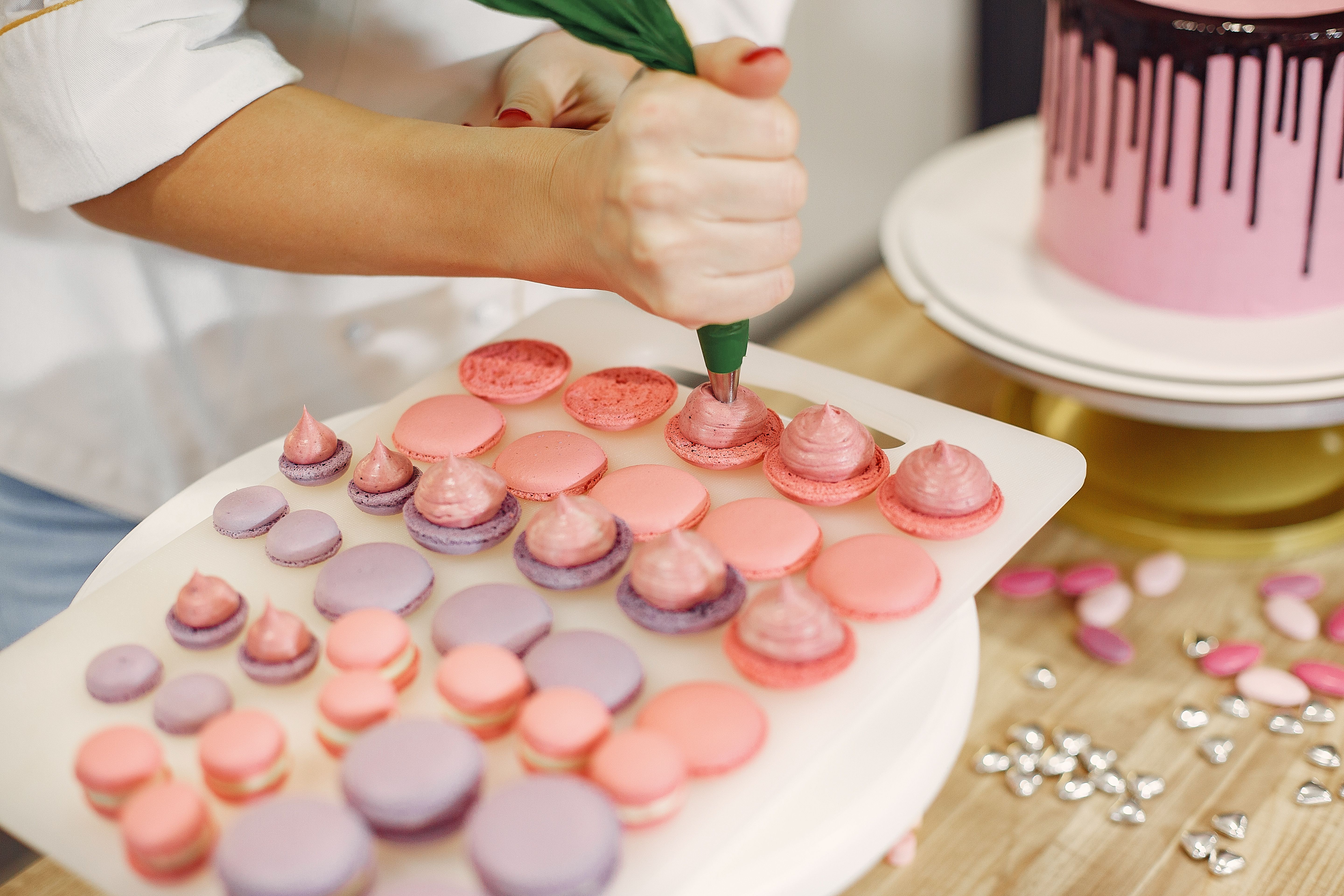 Unrecognizable female confectioner wearing white uniform and piping cream on shells of macaroons from pastry bag  while working in modern kitchen
