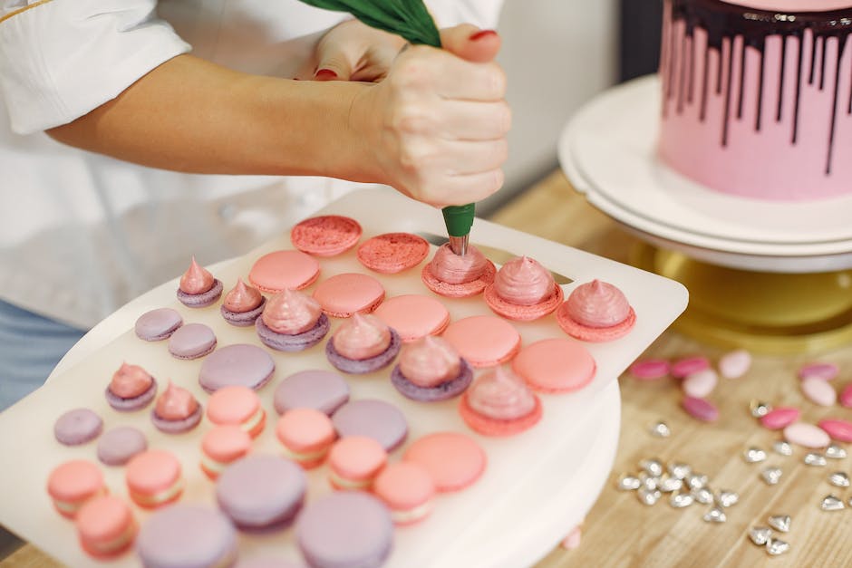 Unrecognizable female confectioner wearing white uniform and piping cream on shells of macaroons from pastry bag while working in modern kitchen