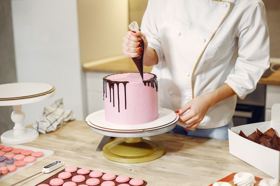 Unrecognizable crop woman in apron making chocolate drips on cake using from pastry bag while preparing pastry in contemporary restaurant