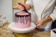 Anonymous female pastry chef in white uniform standing near counter and preparing yummy cake with chocolate drips from pastry bag in kitchen