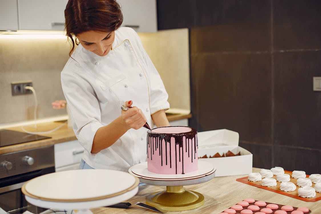 Free Positive female pastry cook in apron standing near counter and making chocolate drips on biscuit cake while preparing dessert in bakery Stock Photo