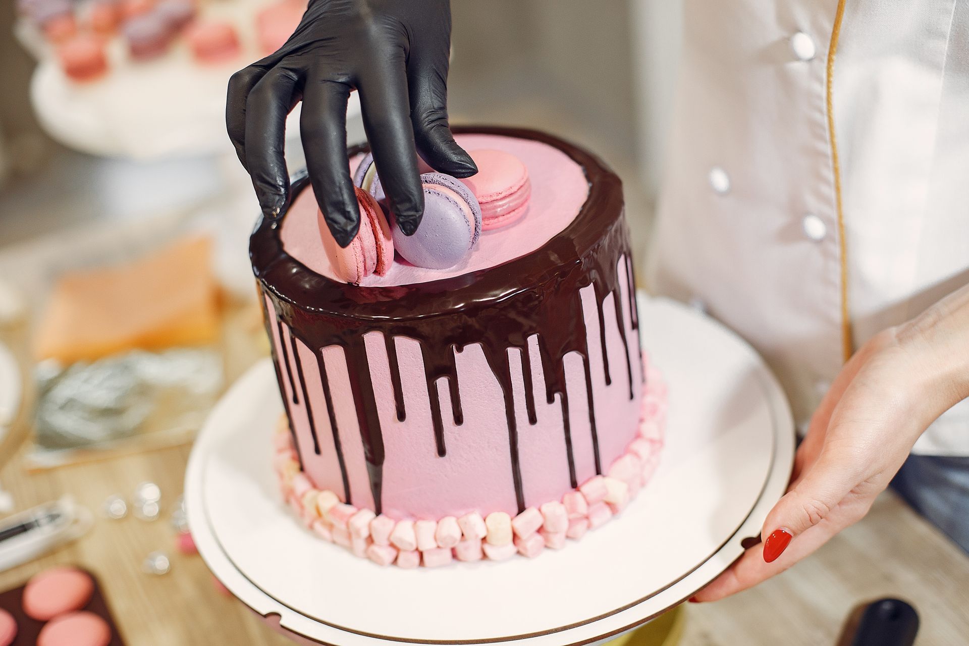From above crop anonymous woman arranging macaroons on top of cake with chocolate drips while working in pastry store
