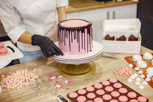 From above crop female cook in uniform and latex gloves placing small sweets on bottom of cake placed on stand near desserts in confectionery
