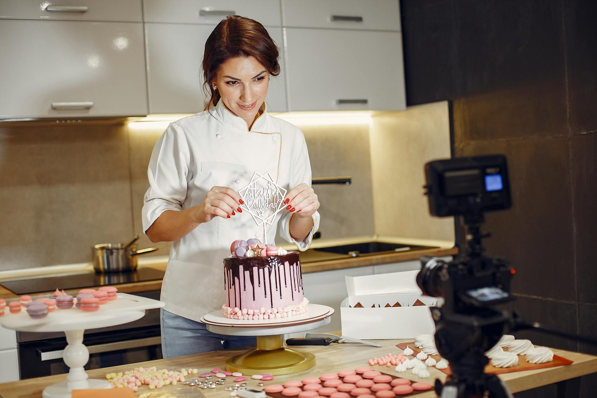 Smiling female chef decorates cake while filming an online baking tutorial in a modern kitchen.