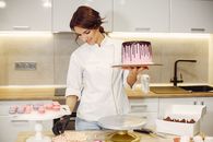 Positive female confectioner in uniform holding pink cake on cardboard with chocolate drips while decorating with colorful marshmallows standing in kitchen