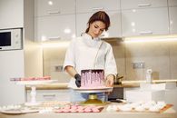 From below of concentrated female pastry cook in uniform garnishing pink cake with marshmallows among confectionery items in kitchen