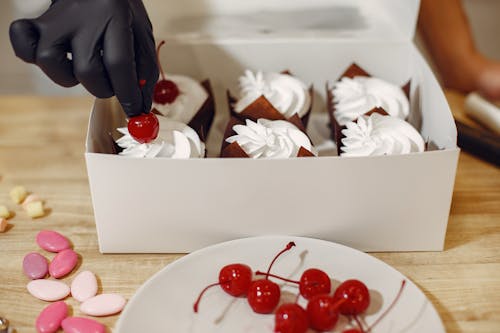 Crop person in black glove decorating cream top of cupcakes with cocktail cherries packing sweets in white box