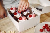 From above of crop anonymous female pastry cook garnishing cream tops of delicious cupcakes with cocktail cherries and macaroons serving in box