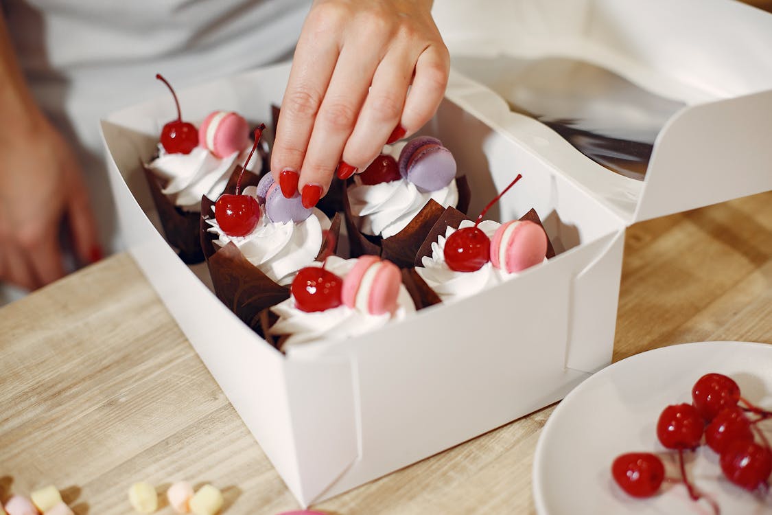 Free From above of crop anonymous female pastry cook garnishing cream tops of delicious cupcakes with cocktail cherries and macaroons serving in box Stock Photo