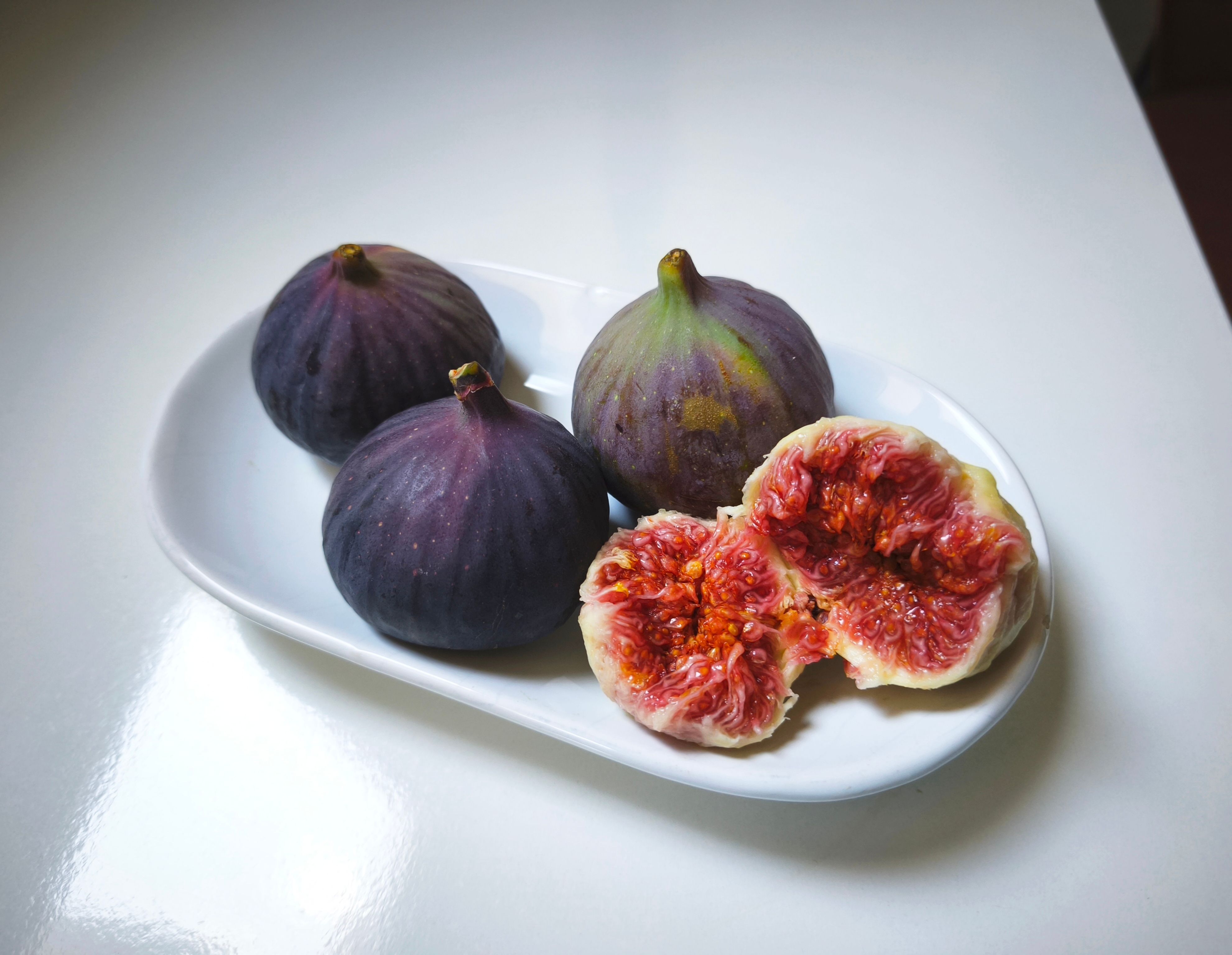 Four ripe figs on a white plate, with one fig halved to show its juicy interior.